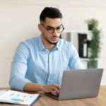 Serious young Arab guy working at his desk with laptop pc, typing on keyboard at office