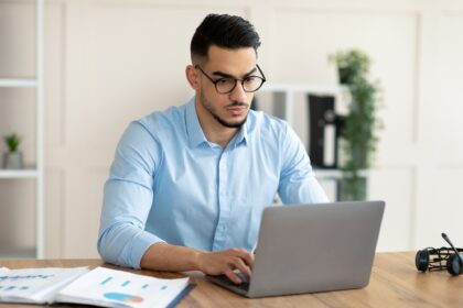 Serious young Arab guy working at his desk with laptop pc, typing on keyboard at office