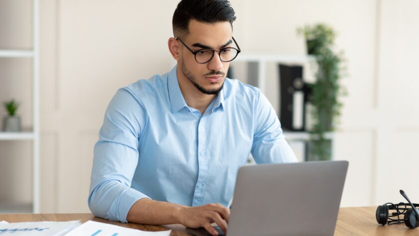 Serious young Arab guy working at his desk with laptop pc, typing on keyboard at office