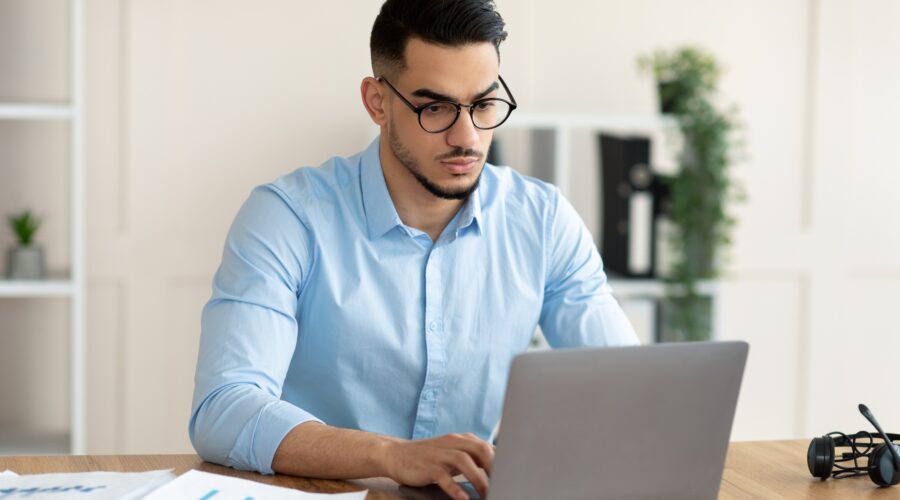 Serious young Arab guy working at his desk with laptop pc, typing on keyboard at office