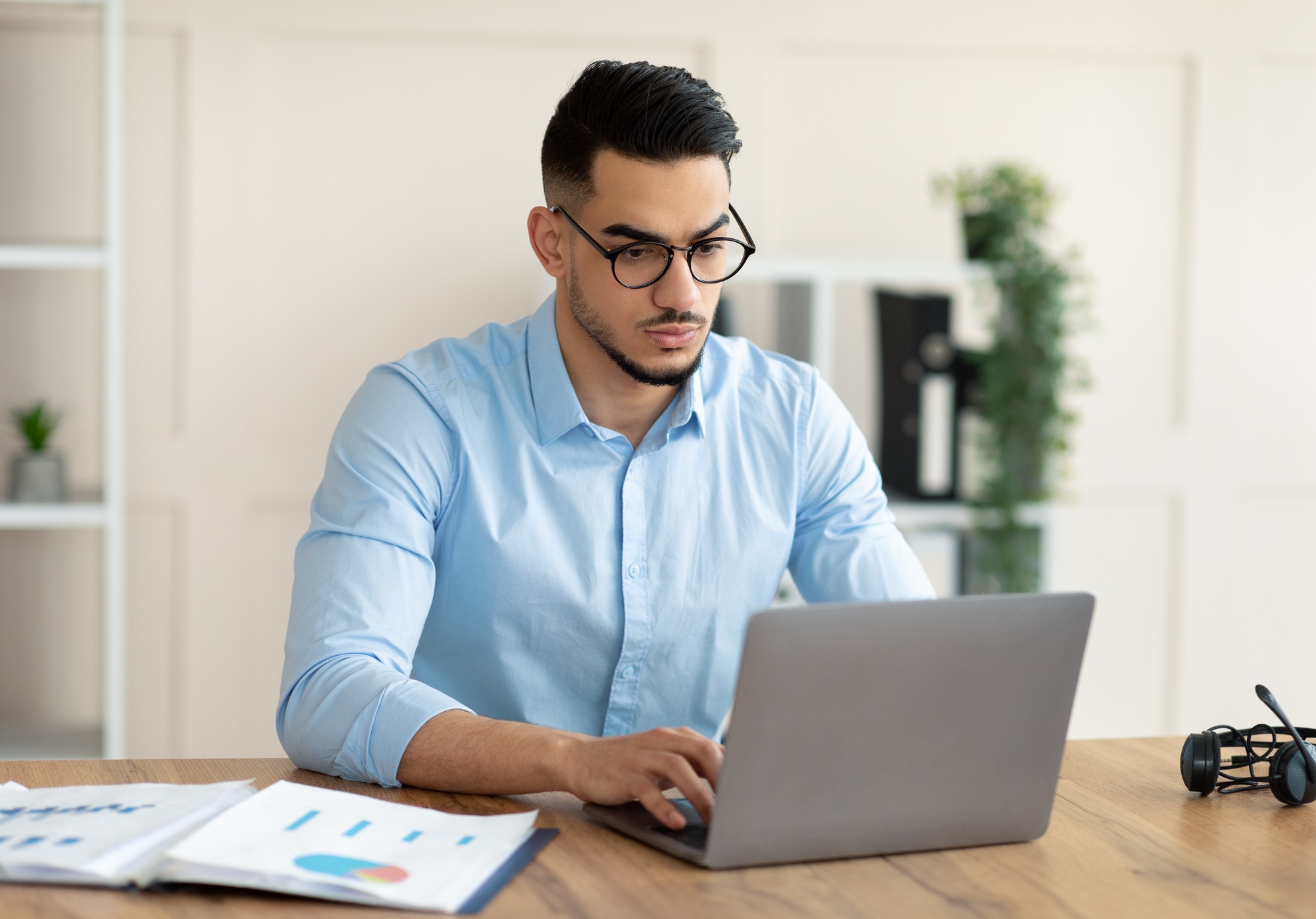Serious young Arab guy working at his desk with laptop pc, typing on keyboard at office
